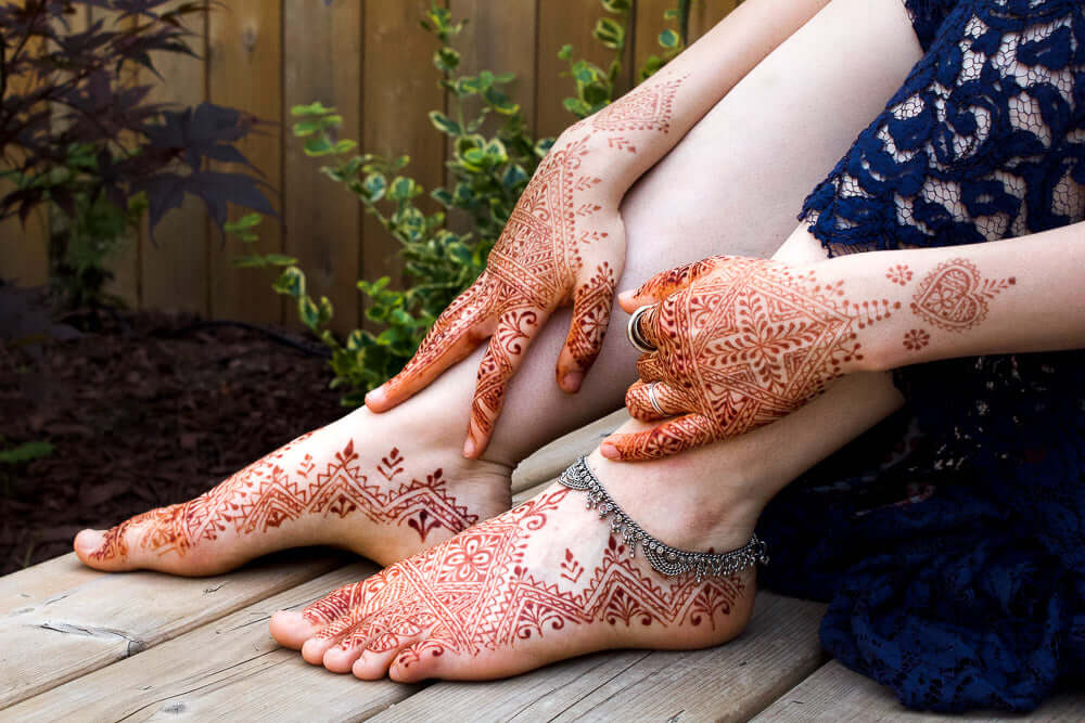 Woman with intricate henna designs on hands and feet sitting on wooden deck.